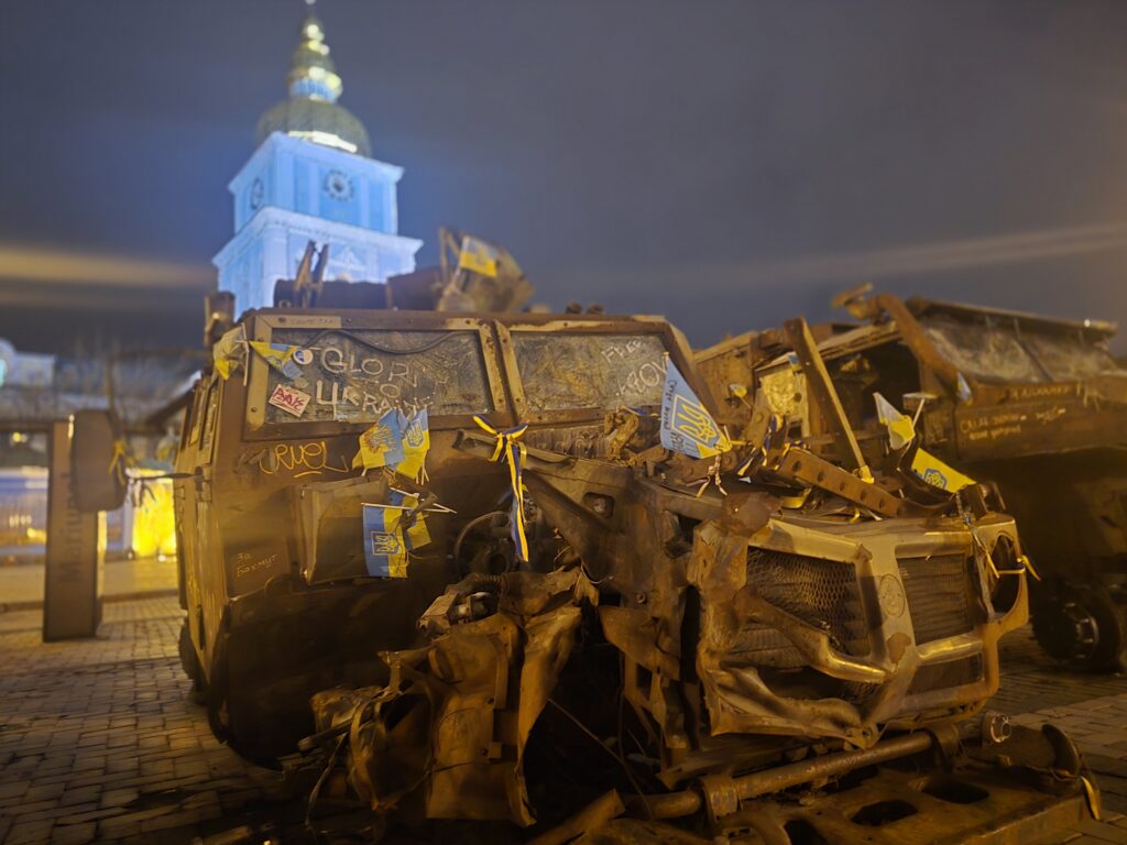 På ett torg i Kyiv står ett förstört ryskt pansarfordon, uppställt som en symbolisk påminnelse om krigets realiteter och Ukrainas motståndskraft. Fordonet är svårt demolerat, dess metall skev och bucklig efter en attack. Det är dekorerat med små ukrainska flaggor och band i gult och blått, som signalerar patriotism och motstånd. På fordonets front och sidor har människor skrivit budskap som "Glory to Ukraine" och andra uttryck för hopp och styrka. I bakgrunden syns en upplyst kyrkobyggnad med ett karakteristiskt guldfärgat torn, vilket förstärker kontrasterna mellan den förstörda krigsmaterielen och den historiska, symboliskt laddade platsen. Kvällen är mörk, men de varma ljusen från torgets belysning och kyrkan skapar en dramatisk stämning.