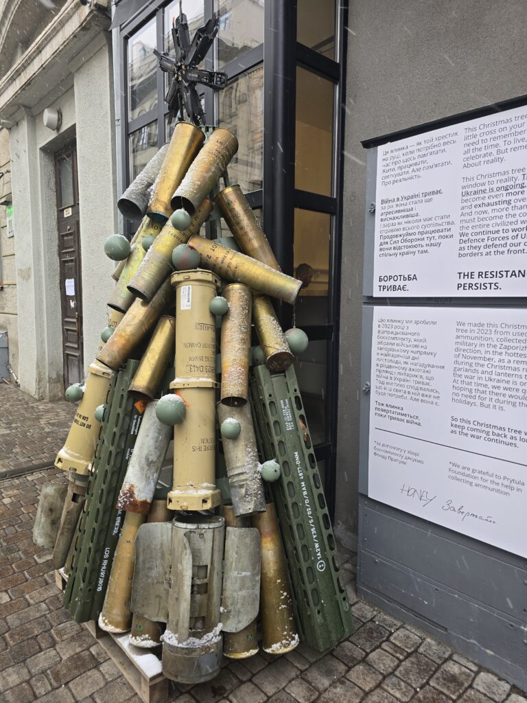 På gatan i Kyiv står en julgran som är allt annat än traditionell – en konstinstallation som kraftfullt symboliserar det pågående kriget. Granen är skapad av använda artillerihylsor, granatdelar och ammunitionstuber. Dess form påminner om en klassisk julgran, men materialen och budskapet är djupt allvarliga. På toppen sitter en stjärna, tillverkad av metall från vapen, vilket förstärker den symboliska laddningen. Vissa av granens delar är dekorerade med gröna kulor, vilket skapar en grotesk kontrast till det material som annars används för destruktion. Bredvid installationen sitter en skylt med text på både ukrainska och engelska. Skylten förklarar installationens betydelse – den är en påminnelse om att julen under ett pågående krig inte är som vanliga jular. Texten beskriver att detta träd är tillägnat de liv som har gått förlorade och den kamp som fortsätter för Ukrainas frihet och överlevnad. Budskapet understryker vikten av att komma ihåg verkligheten bortom firandets yta och att förstå krigets påverkan på samhället. Marken runt granen är täckt av ett tunt lager snö, vilket ger en känsla av vinter och förstärker installationens visuella intryck. Byggnaden bakom är modern och enkel, vilket ger en neutral bakgrund som låter granen stå i fokus.